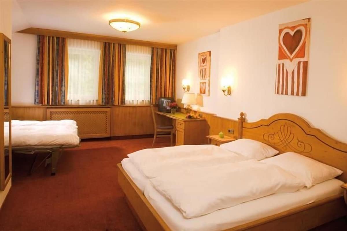Une chambre d’hôtel avec un grand lit, un bureau avec une chaise, une fenêtre avec des rideaux et un mur orné d’un décor en forme de cœur.