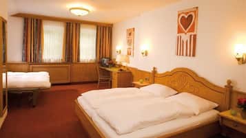A hotel room with a large bed, a desk with a chair, a small table with a lamp, and a window with curtains.