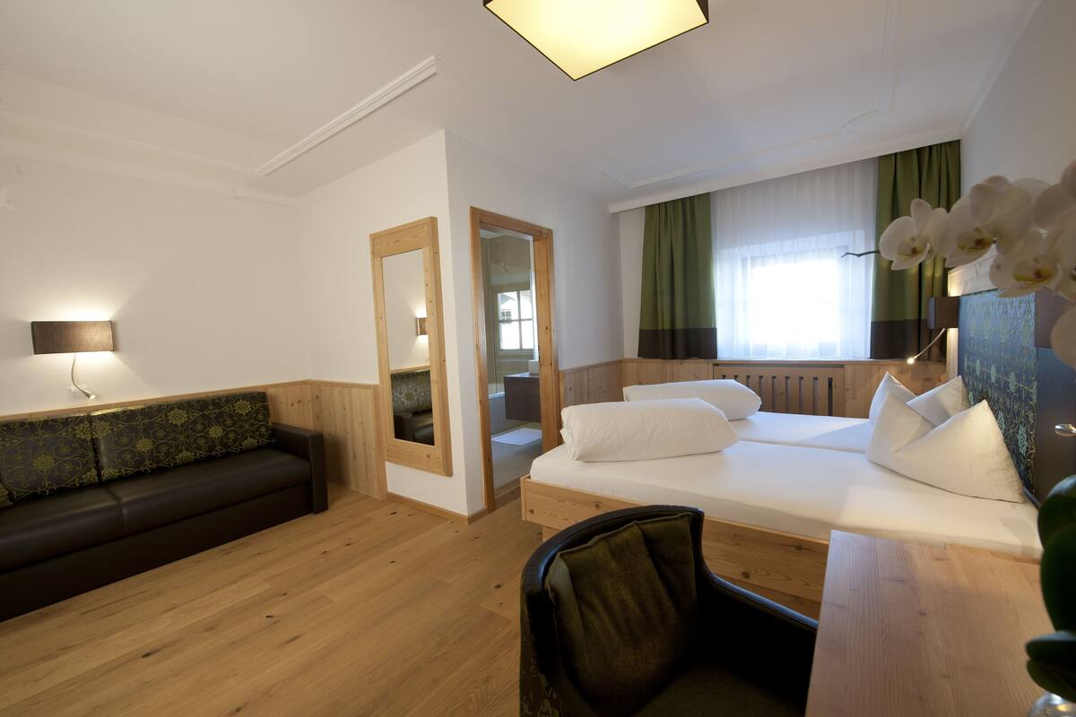 Une chambre d’hôtel avec un plancher en bois, un lit avec du linge de lit blanc, un canapé vert foncé, un bureau en bois et un miroir.