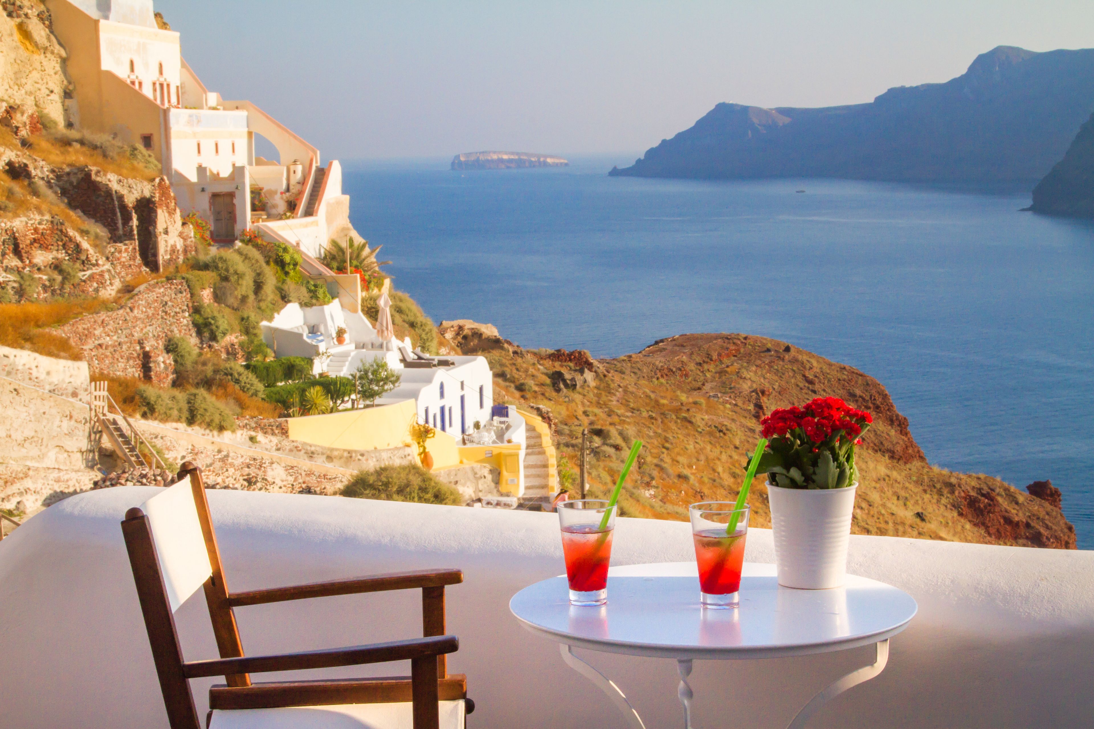 Una mesa con dos bebidas y una flor, con vistas a un paisaje costero con edificios y el mar.