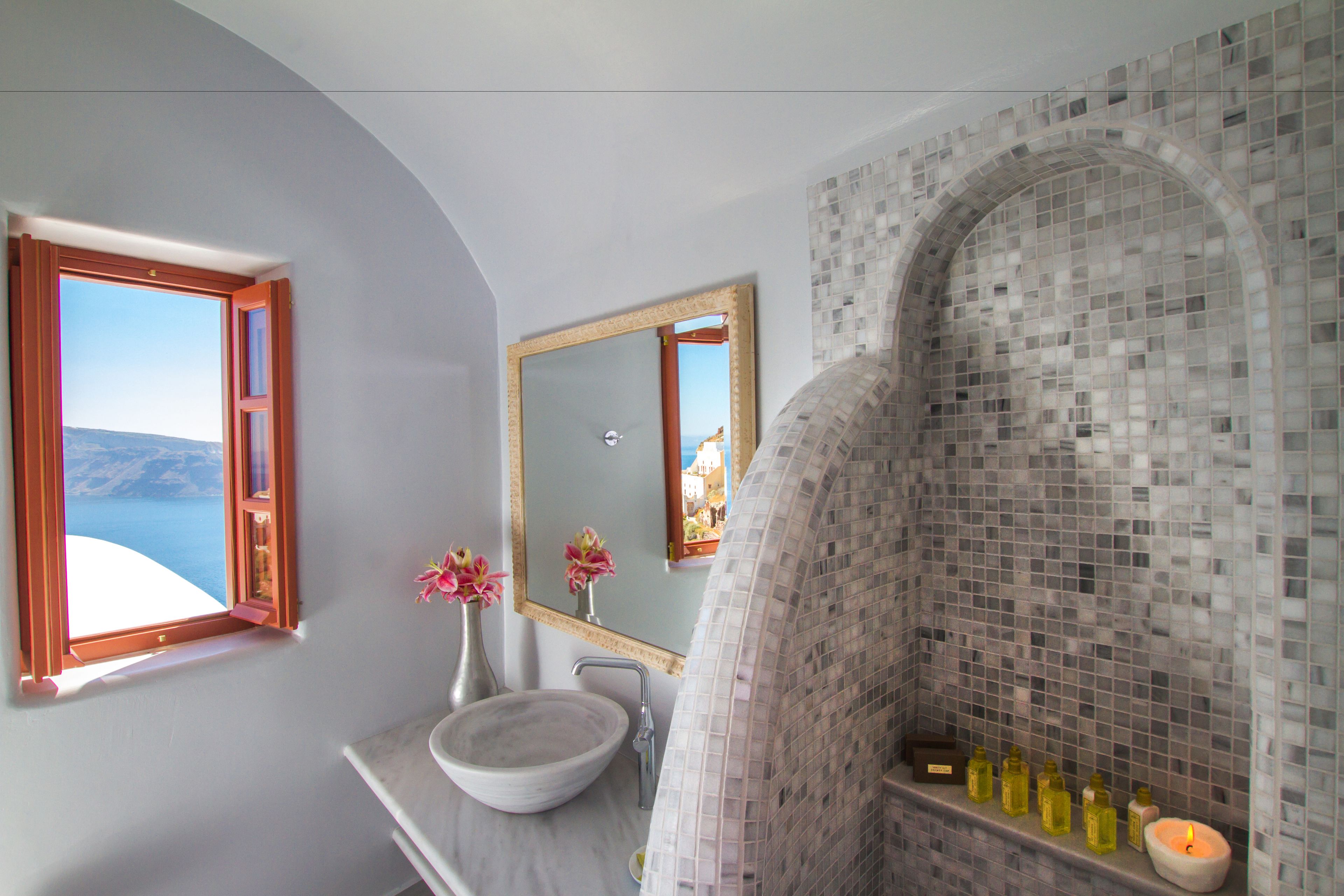 A bathroom with a large arched mirror, a white sink, and a view of the sea through a window.
