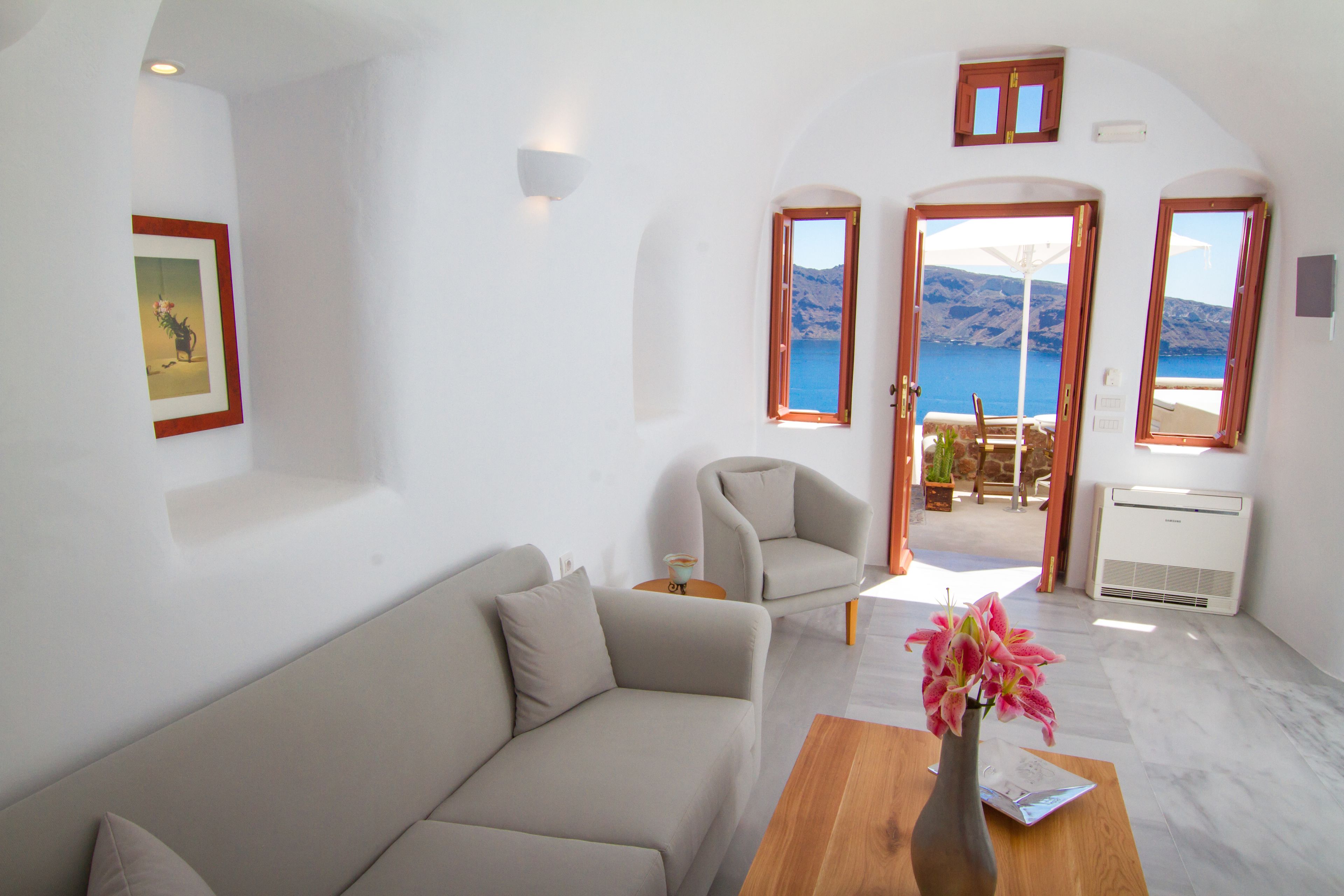 A modern living room with a sofa, armchair, and a wooden coffee table with a vase of flowers. There are two doors leading to a balcony with a view of the sea and mountains.