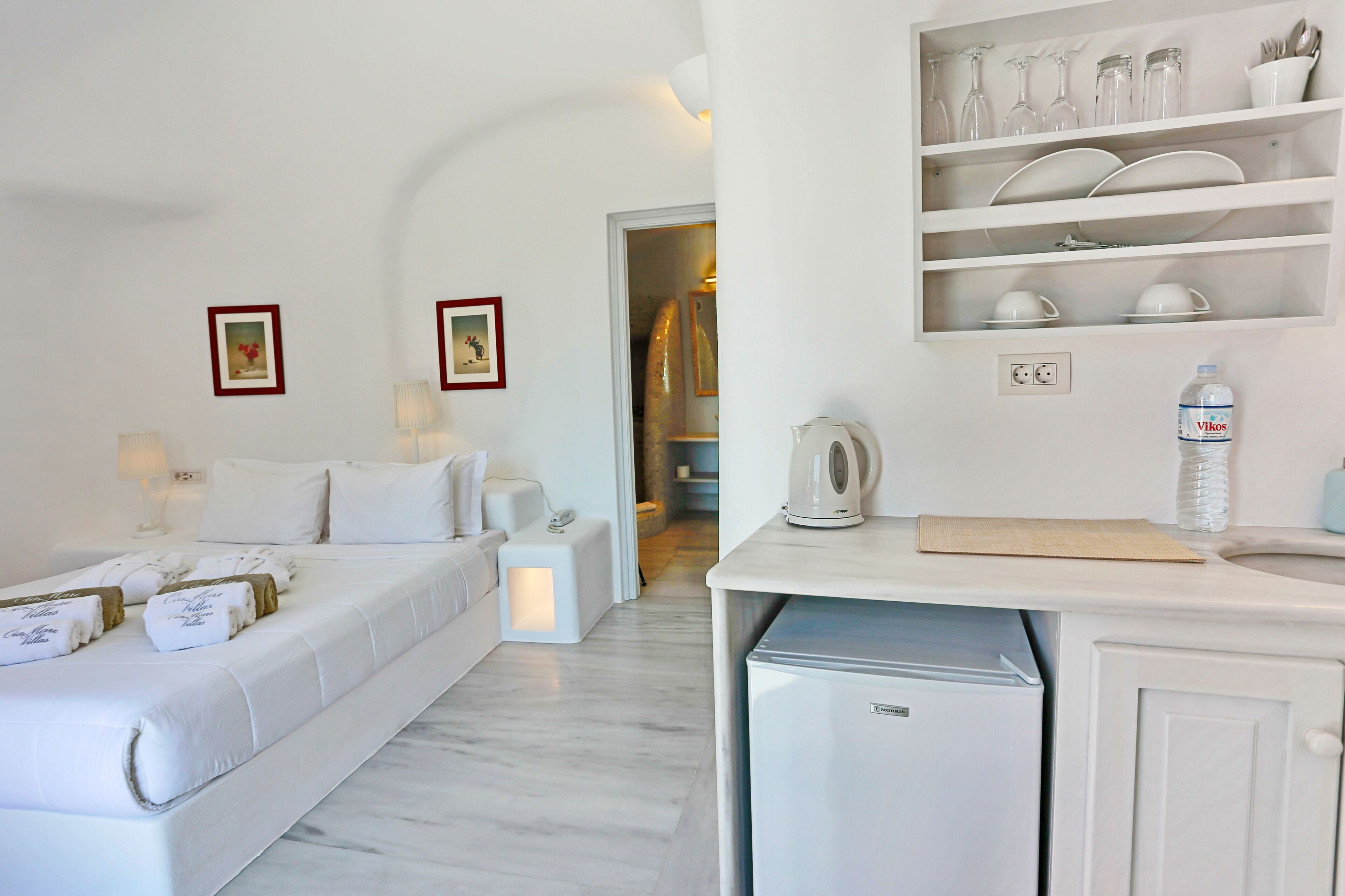Habitación de hotel con cama, lámparas de noche, una mesita con hervidor, un mini refrigerador y una repisa con cristalería y vajilla.
