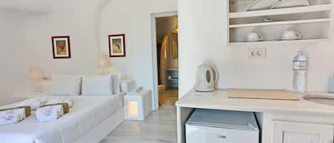 Habitación de hotel con cama, lámparas de noche, una mesita con hervidor, un mini refrigerador y una repisa con cristalería y vajilla.