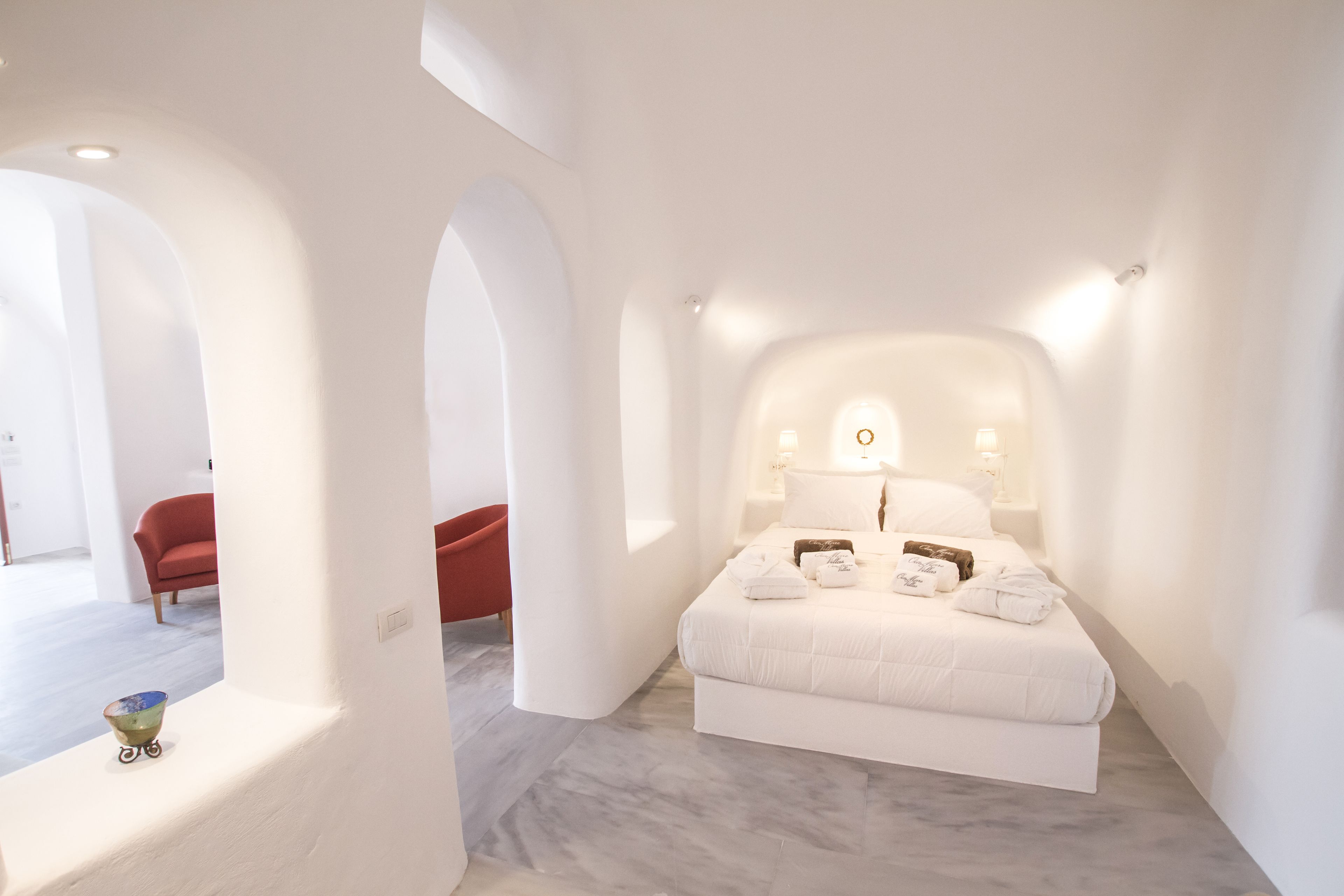 A modern bedroom with a large bed, white walls, and a marble floor. There is a red chair in the background and a small table with a bowl.