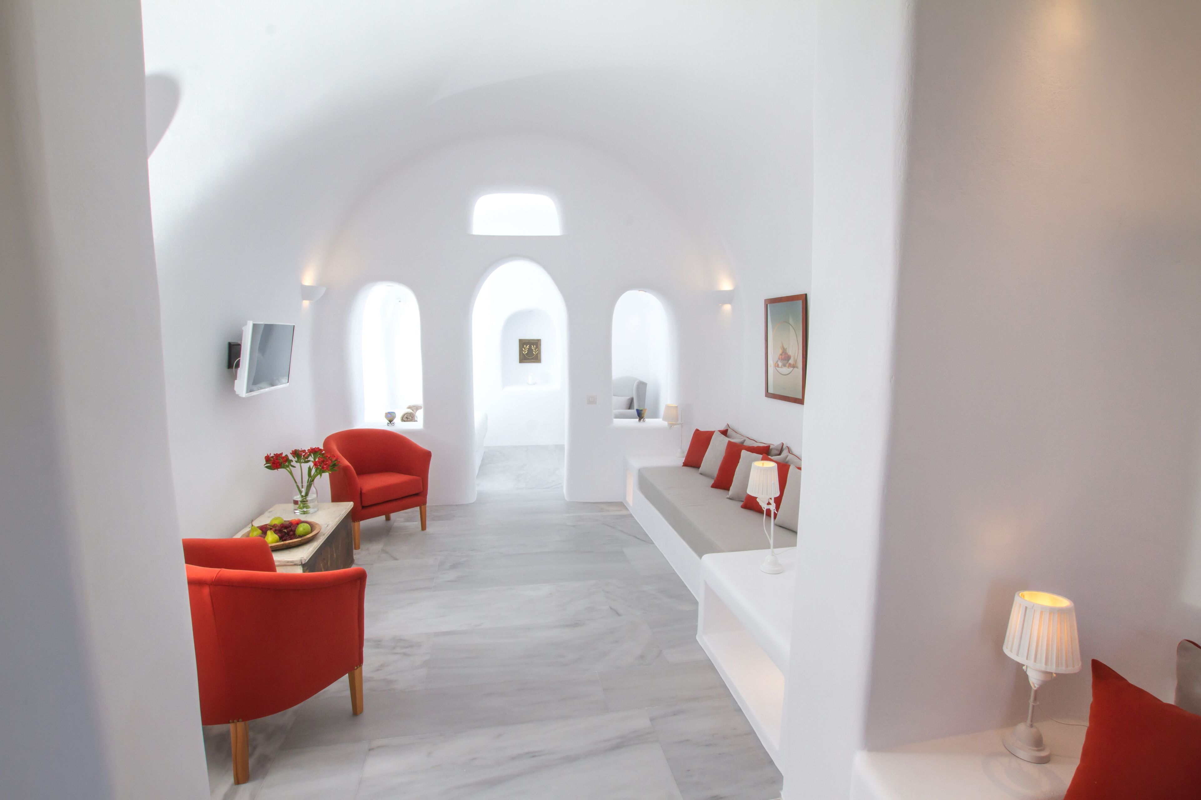 Um interior moderno com esquema de cores branco, com área de jantar com cadeiras vermelhas, um sofá com almofadas vermelhas e uma TV fixada na parede.