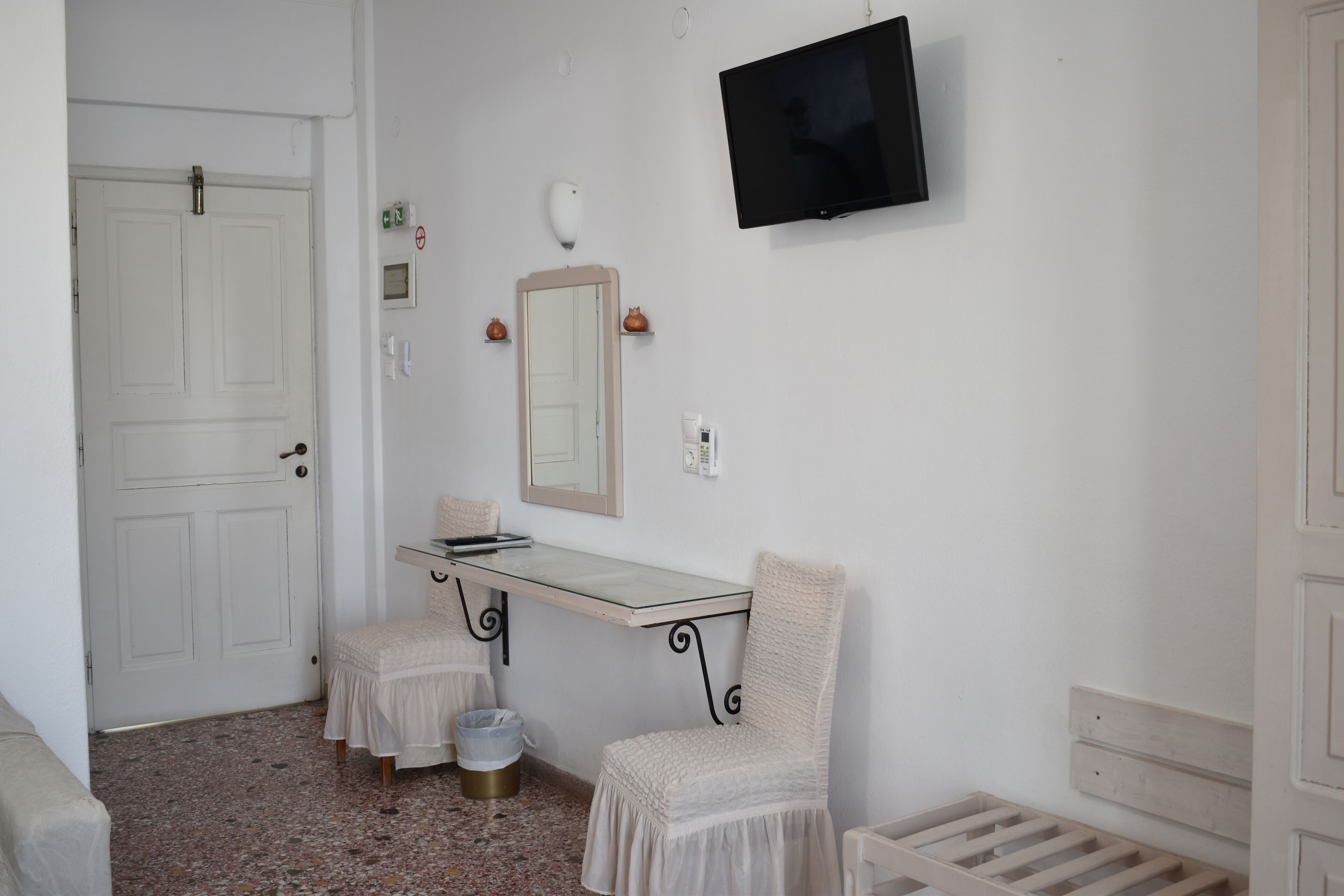 A small, clean room with a white door, a TV mounted on the wall, a small table with a chair, and a trash bin.