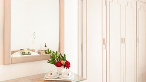 Une chambre d’hôtel avec un grand miroir, un lit agrémenté d’un coussin décoratif, une petite table sur laquelle est posé un vase de fleurs et une bouteille de vin.