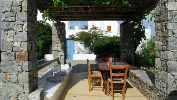 Un espace extérieur aménagé avec des murs en pierre, comprenant une table et des chaises, une pergola au-dessus et une vue sur un bâtiment blanc agrémenté de touches de bleu.