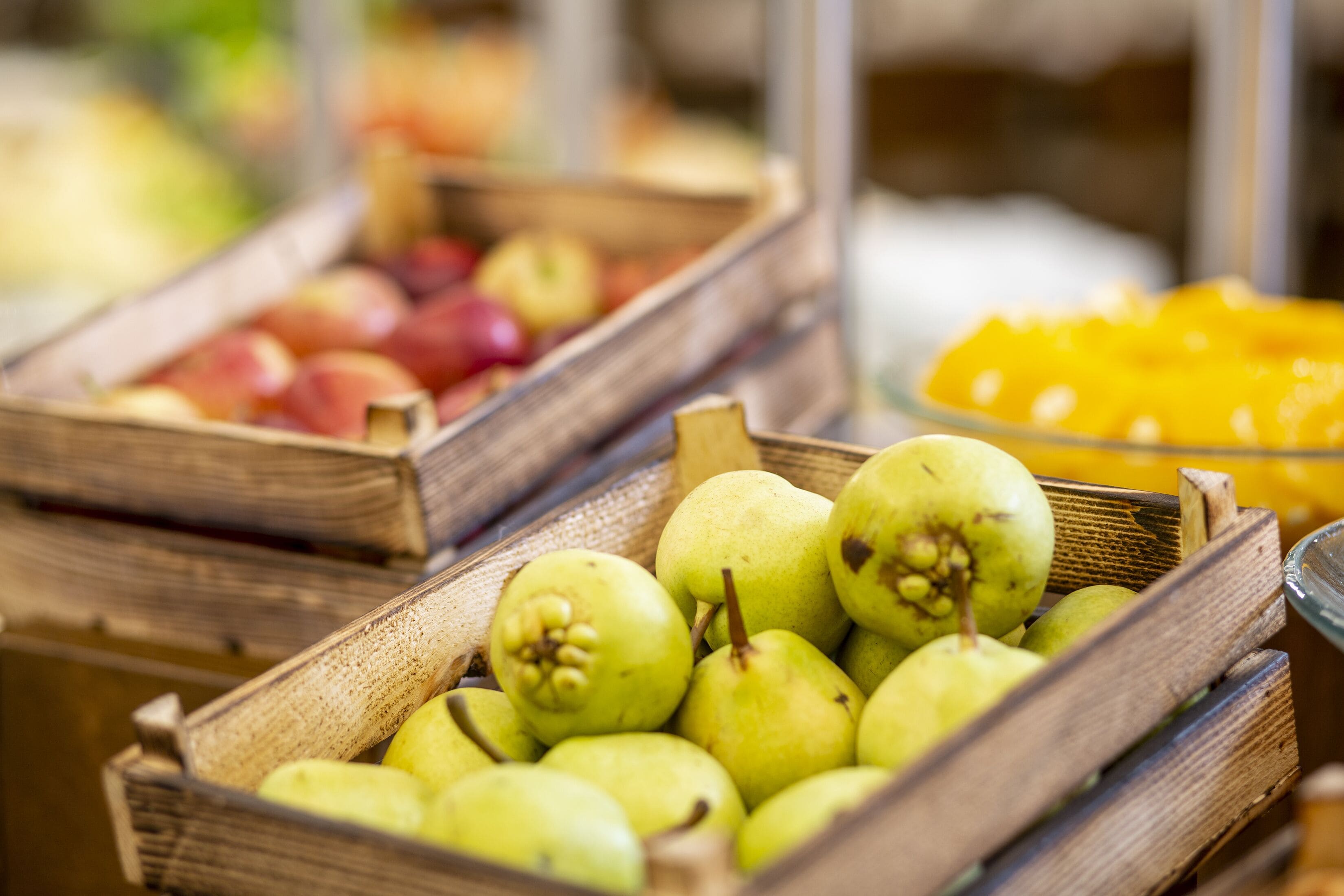 A wooden crate filled with green pears, a wooden crate with red apples, and a glass bowl with yellow fruit.