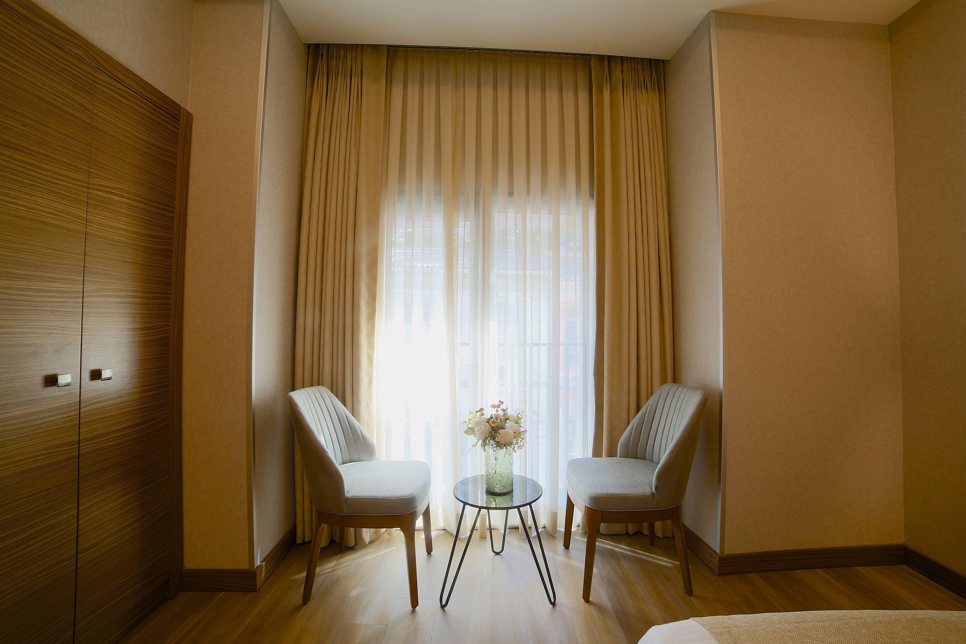 Um quarto com duas cadeiras, uma mesinha com um vaso, e uma janela grande com cortinas.