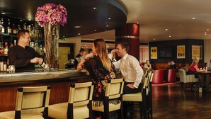 Zona de bar con un cantinero, una mujer con vestido floral y un hombre con camisa blanca. Hay varias sillas y mesas, y un jarrón grande con flores en la barra.