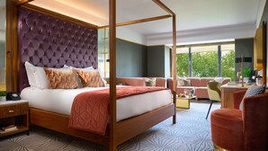 Una cama con dosel, un cabecero capitonado, un sofá, una silla y un escritorio en una habitación de hotel moderna con ventanas amplias.