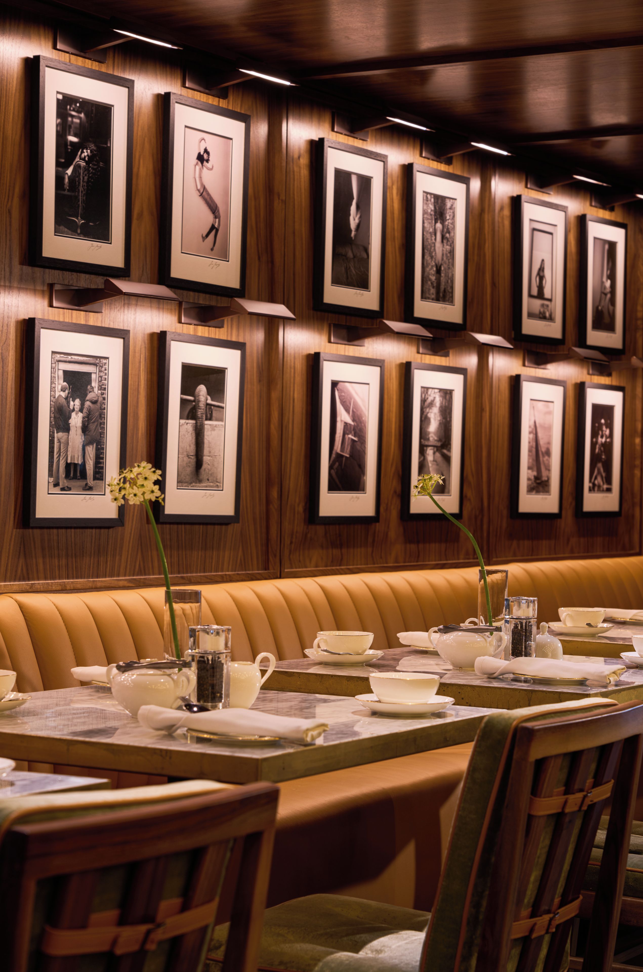 Interior de un restaurante con un largo banco de madera, fotografías en blanco y negro enmarcadas en la pared y una mesa dispuesta con vajilla blanca y un florero.