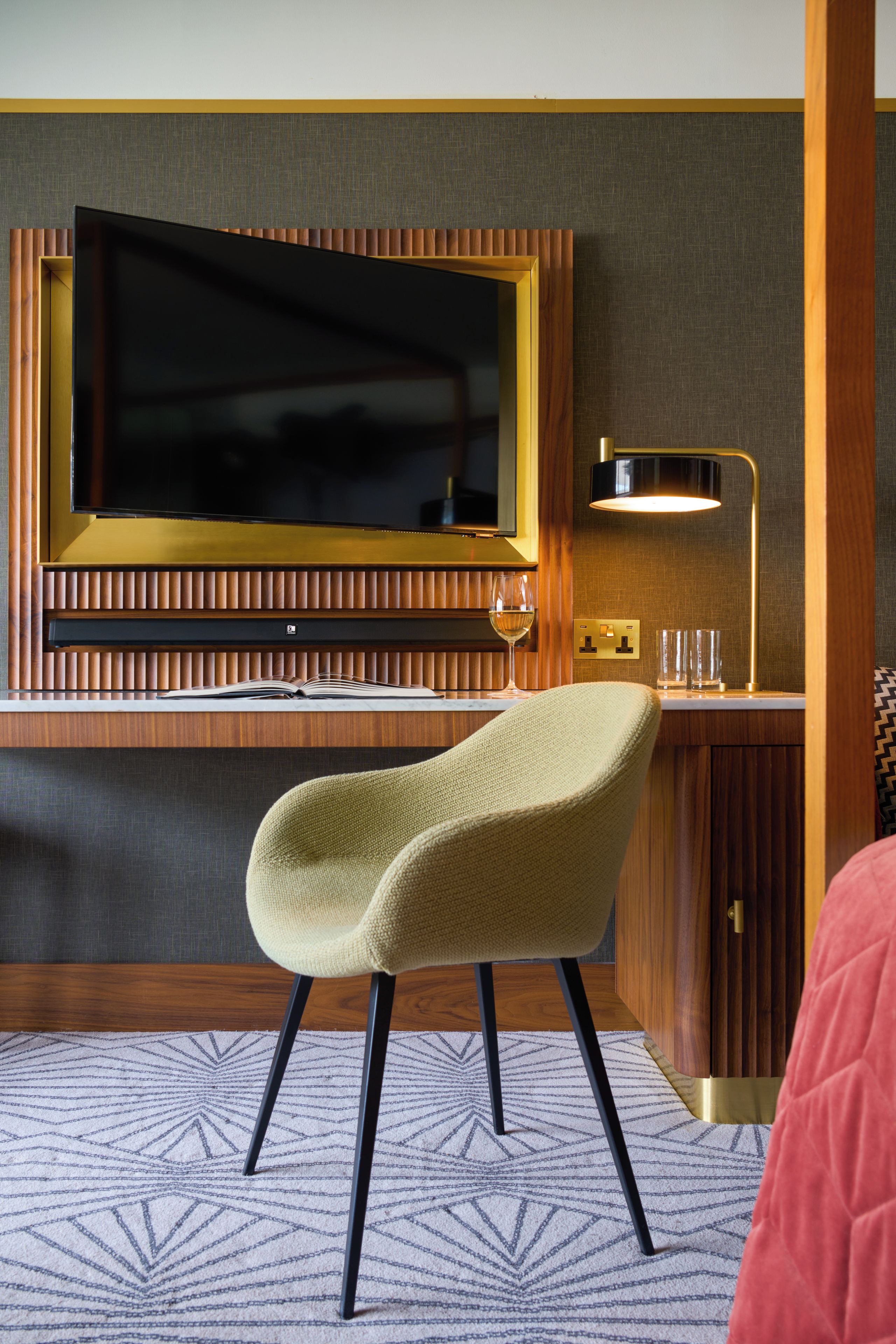 A hotel room with a flat-screen TV mounted on a wooden panel wall, a desk with a lamp, a green chair, and a patterned rug.
