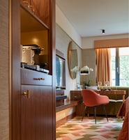A modern room with a wooden wardrobe, a large mirror, a pink chair, a dining table with a vase of flowers, and a window with curtains.