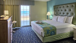 Quarto de hotel com cama, cômoda, mesa de cabeceira, luminária e vista para o oceano através de uma janela grande.