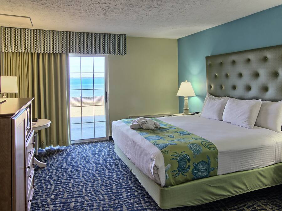 Una camera d'hotel con un letto, una cassettiera, un comodino, una lampada e vista sull'oceano attraverso un'ampia finestra.