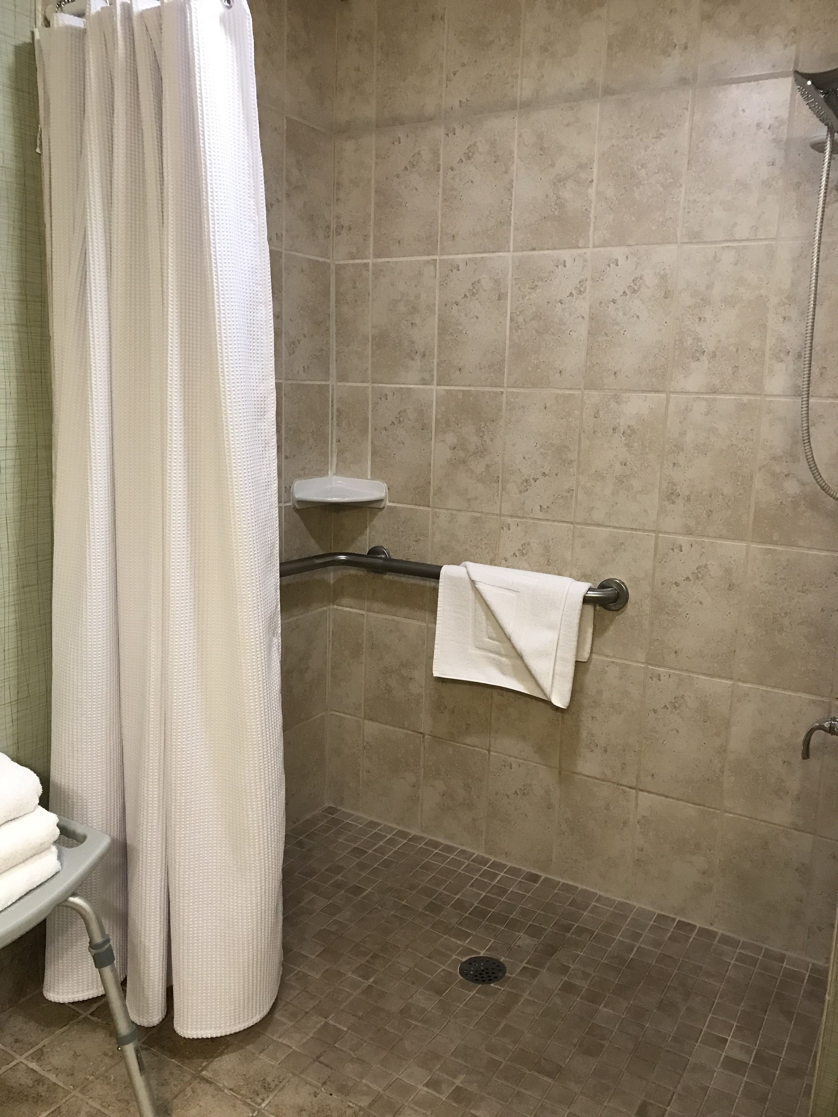 A shower area with a white shower curtain, a handheld showerhead, a grab bar, a small white shelf, and a towel rack with a towel.