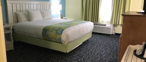 Ein Hotelzimmer mit Bett, Nachttisch, Schreibtisch, Stuhl, einem Fenster mit Vorhängen und einem Wandheizkörper.