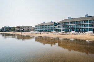 Un hotel frente a la playa con varios balcones, una playa de arena y una masa de agua tranquila al frente.