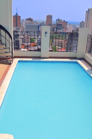 A rooftop swimming pool with a city view, surrounded by buildings and a metal railing.