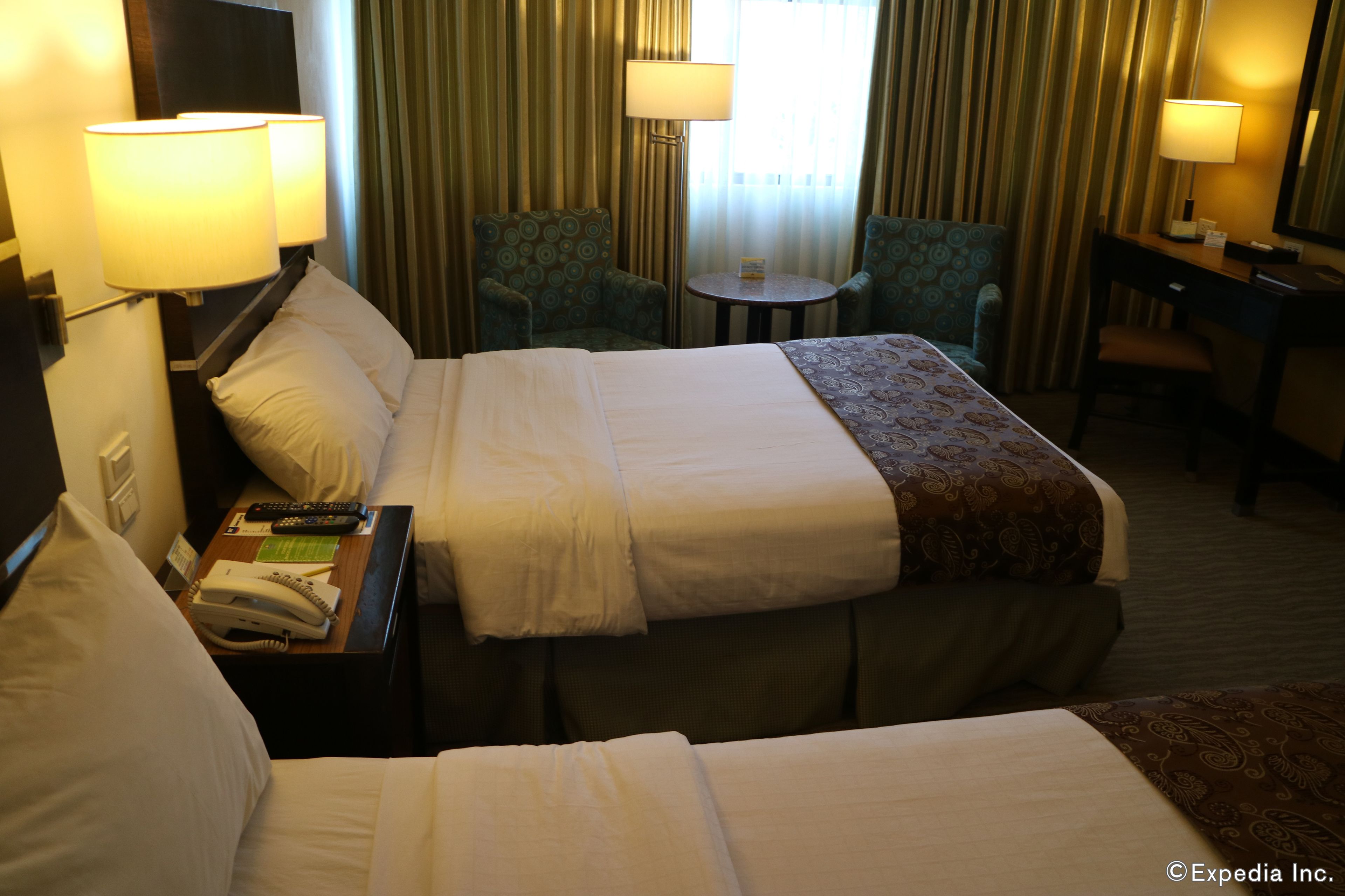 Hotellihuone, jossa on suuri sänky, kaksi nojatuolia, pieni pöytä ja työpöytä, jossa on lamppu.