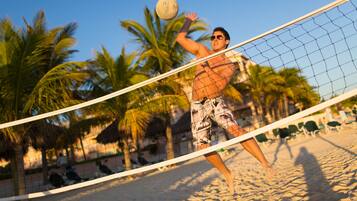 Partido de voleibol de playa con palmeras y sillas de playa al fondo.