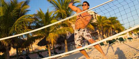 A beach volleyball game with palm trees and beach chairs in the background.