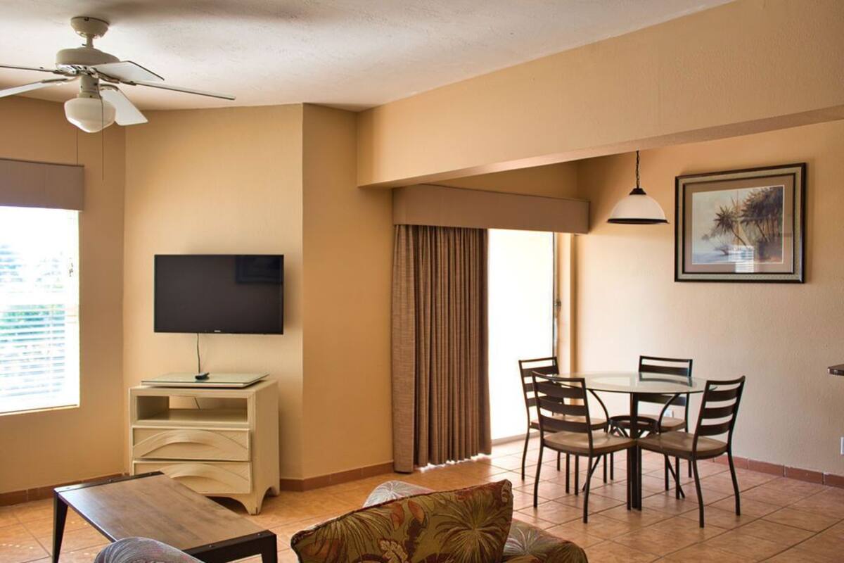 A living room with a TV, a dining table with chairs, a ceiling fan, and a window with curtains.