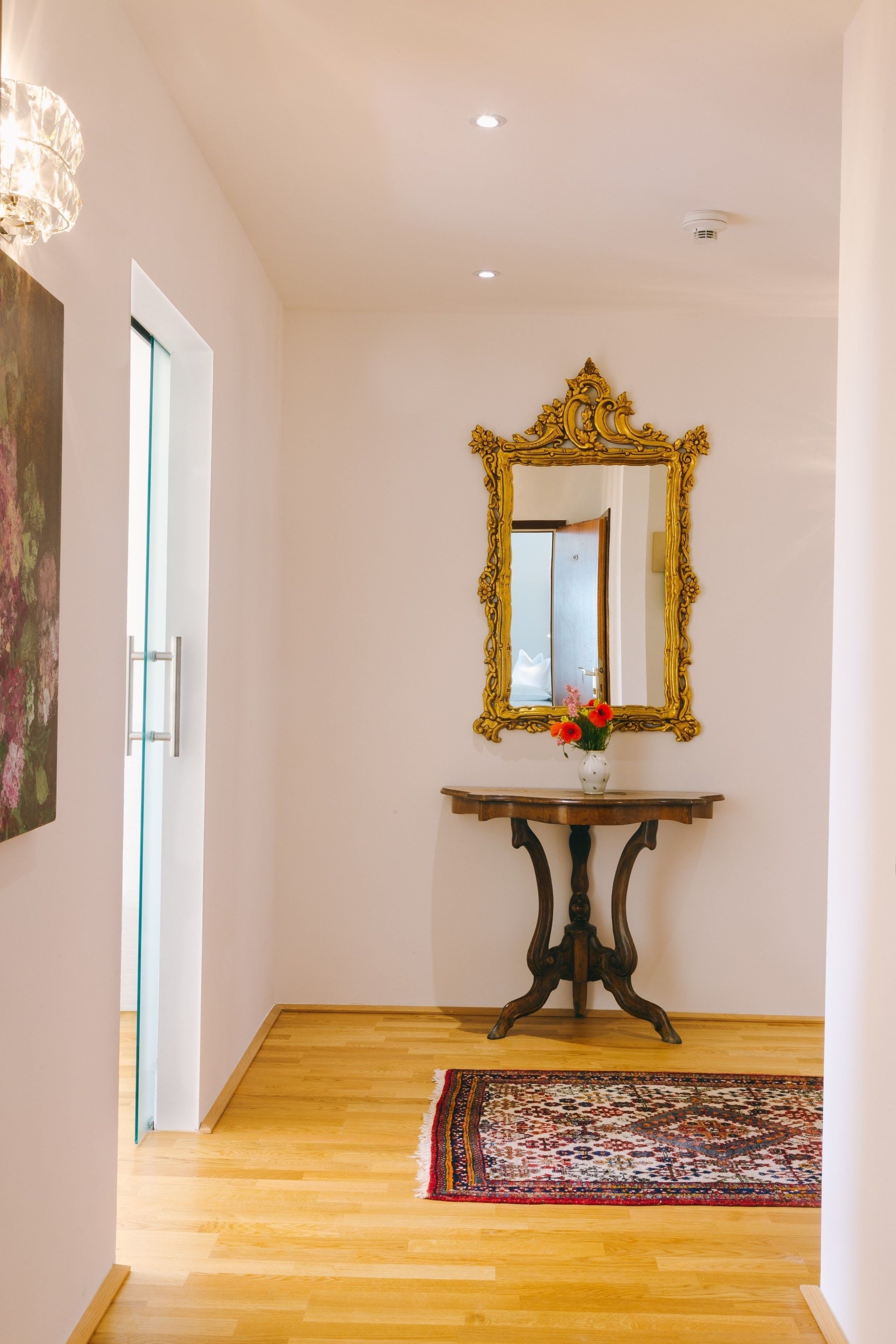 A hallway with a wooden floor, a decorative mirror, a vase with flowers, and a patterned rug.