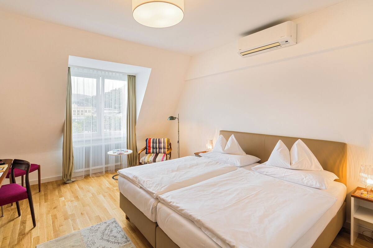 Ein Hotelzimmer mit zwei Betten, einem kleinen Tisch, einem Sessel, einem Fenster mit Vorhängen und einer Klimaanlage.