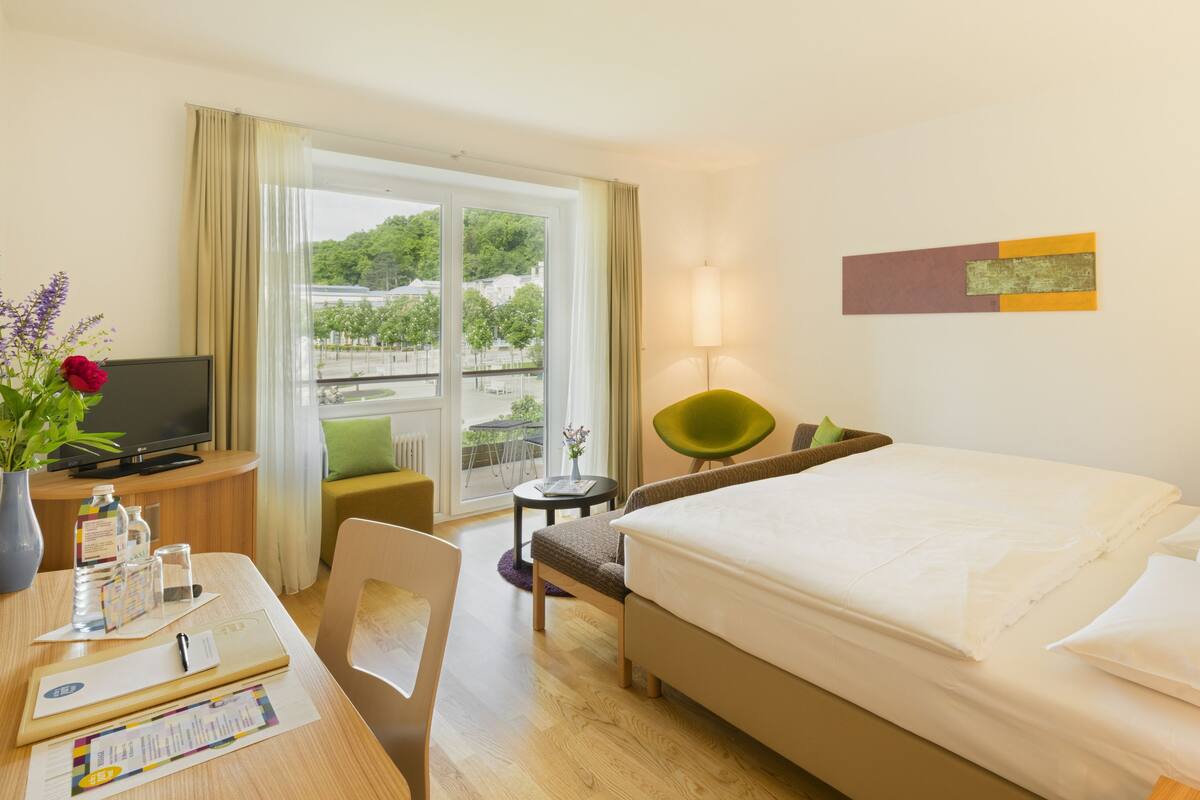 Ein Hotelzimmer mit Bett, Schreibtisch mit Computer, Stuhl, kleinem Tisch mit Lampe und Fenster mit Blick auf Bäume und Gebäude.