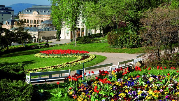 A park with benches, a flower garden, and a building with a dome and arched windows. - At the Park Hotel (Baden)