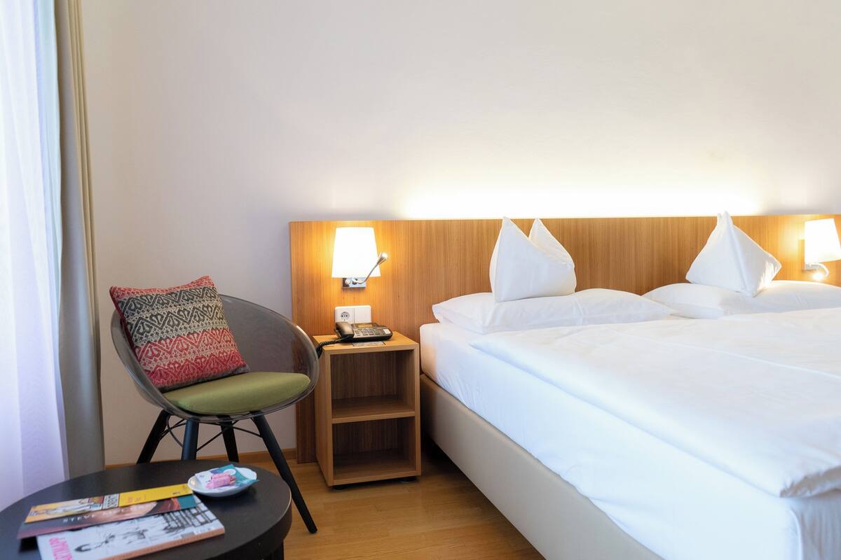 Ein Hotelzimmer mit Bett, Nachttisch, Stuhl und einem kleinen Holfnachtkästchen mit Lampe.