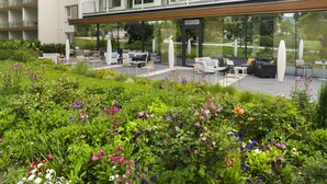 A modern multi-story building with a glass facade, outdoor seating area, and a garden with various flowers. - At the Park Hotel (Baden)