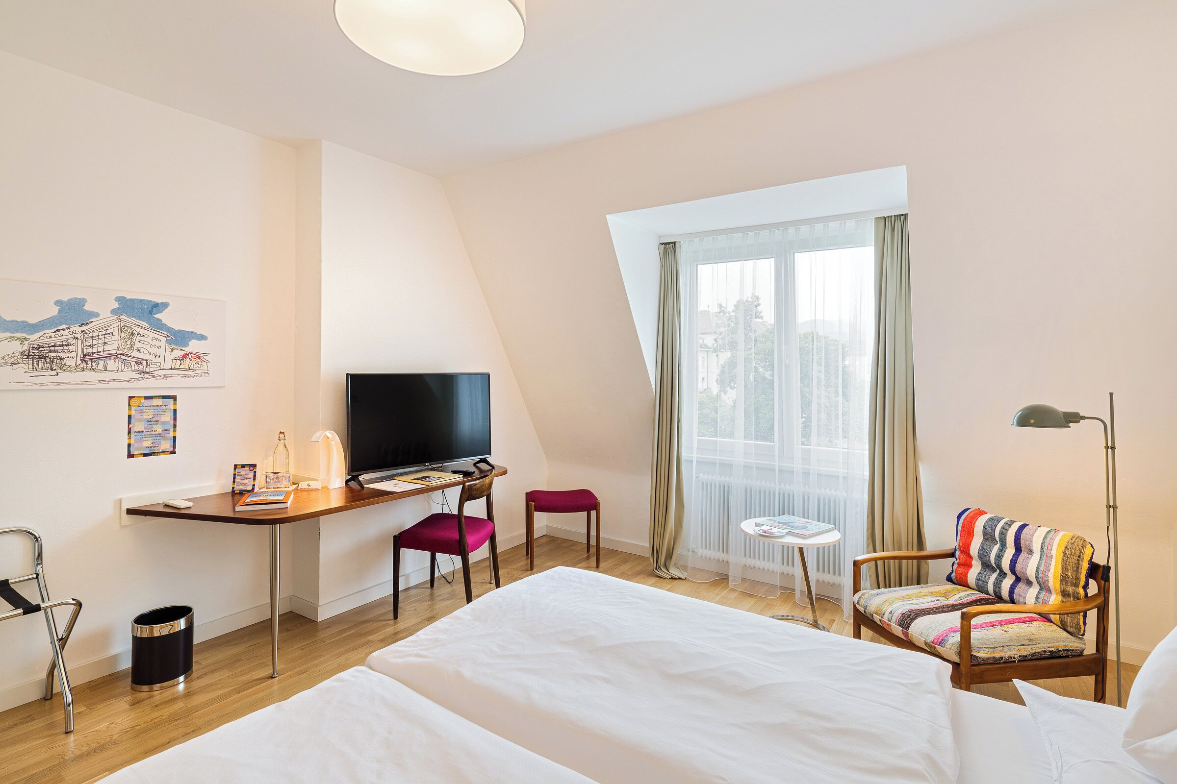 Ein Hotelzimmer mit Bett, Schreibtisch, Stuhl, Fernseher und einem Fenster mit Vorhängen.