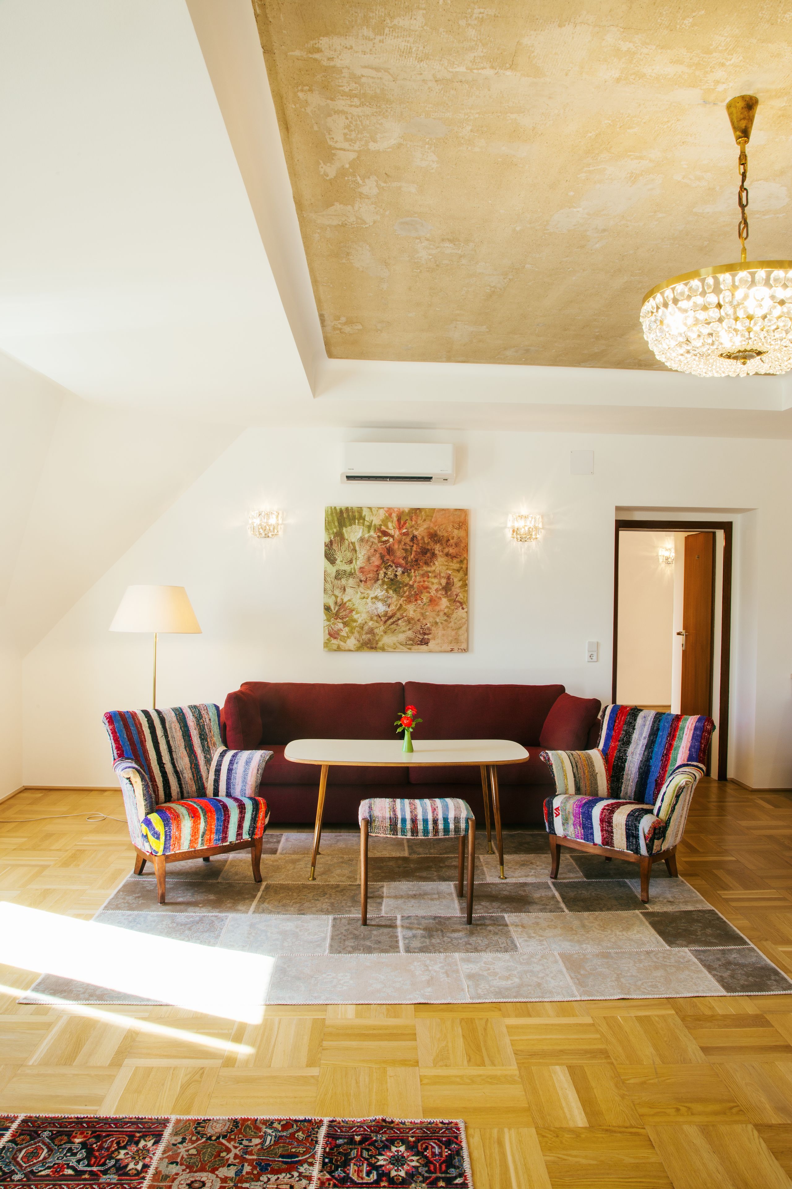Ein gemütliches Wohnzimmer mit einem roten Sofa, zwei gestreiften Sesseln, einem kleinen Tisch mit einer Vase und einer Kristallleuchter.