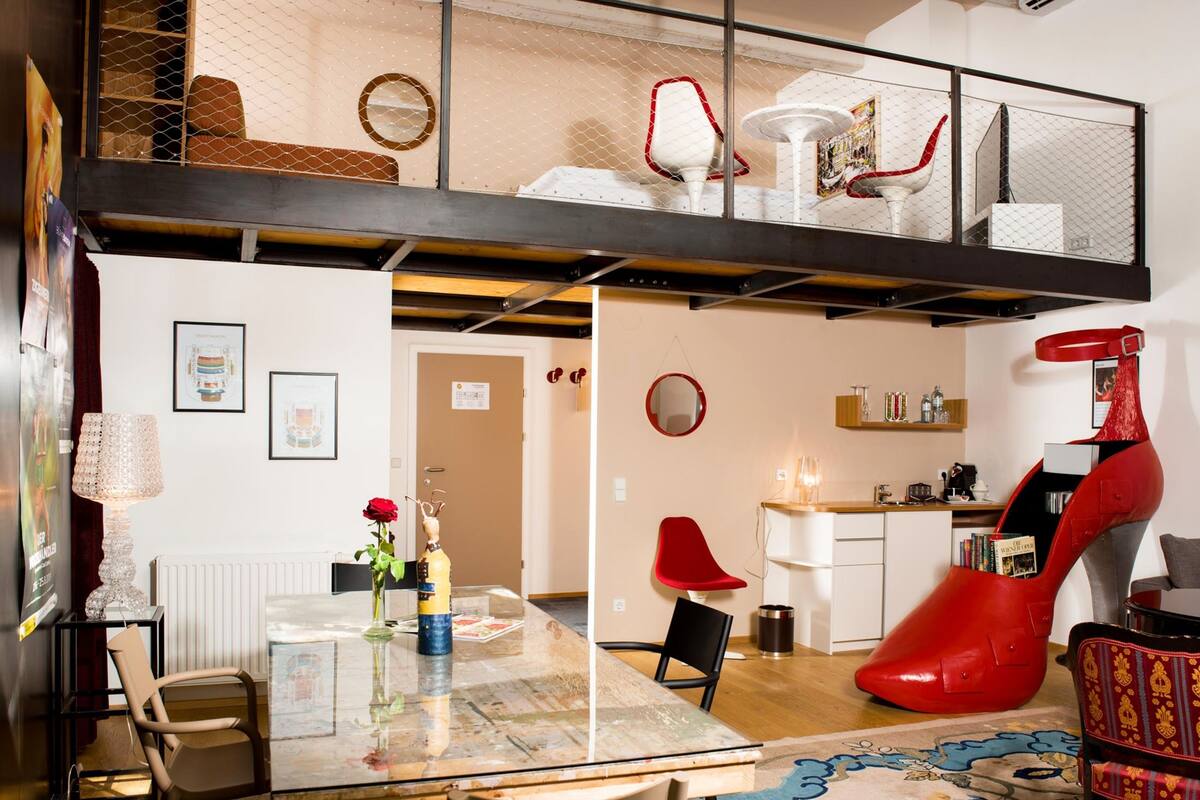 Ein modernes Loft-Apartment mit einem Glastisch, roten Stühlen, einer Küchenzeile und einem ungewöhnlichen, roten High-Heel-Schuh als Sitzgelegenheit.