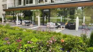A modern building with a cafe-bar on the ground floor, outdoor seating with white furniture, and a well-maintained garden with various plants and flowers. - At the Park Hotel (Baden)