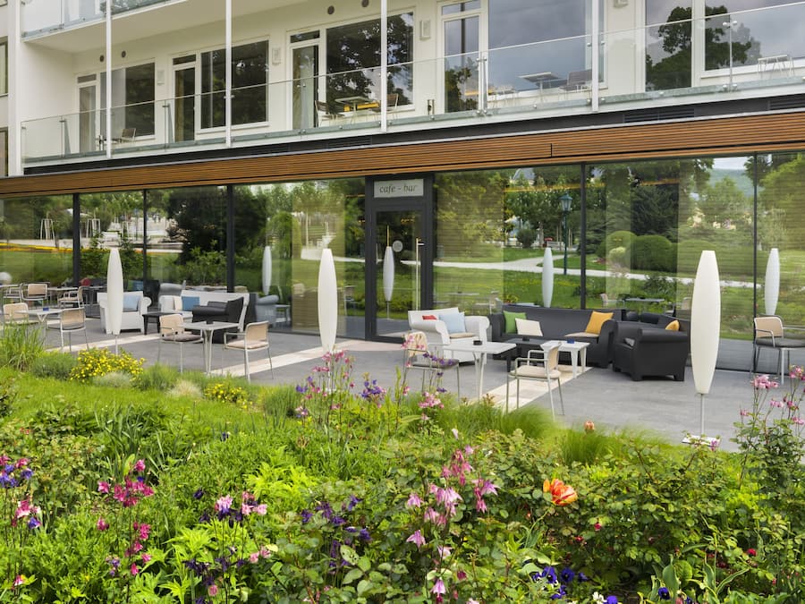 A modern building with a cafe-bar on the ground floor, outdoor seating with white furniture, and a well-maintained garden with various plants and flowers.