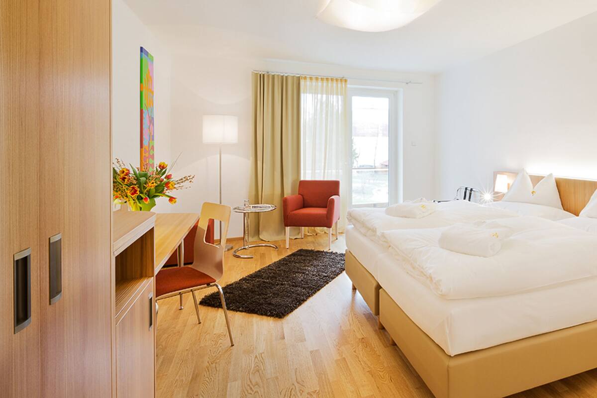 Ein modernes Hotelzimmer mit einem großen Bett, einem Schreibtisch, einem Sessel, einem kleinen Tisch und einem Fenster mit Vorhängen.