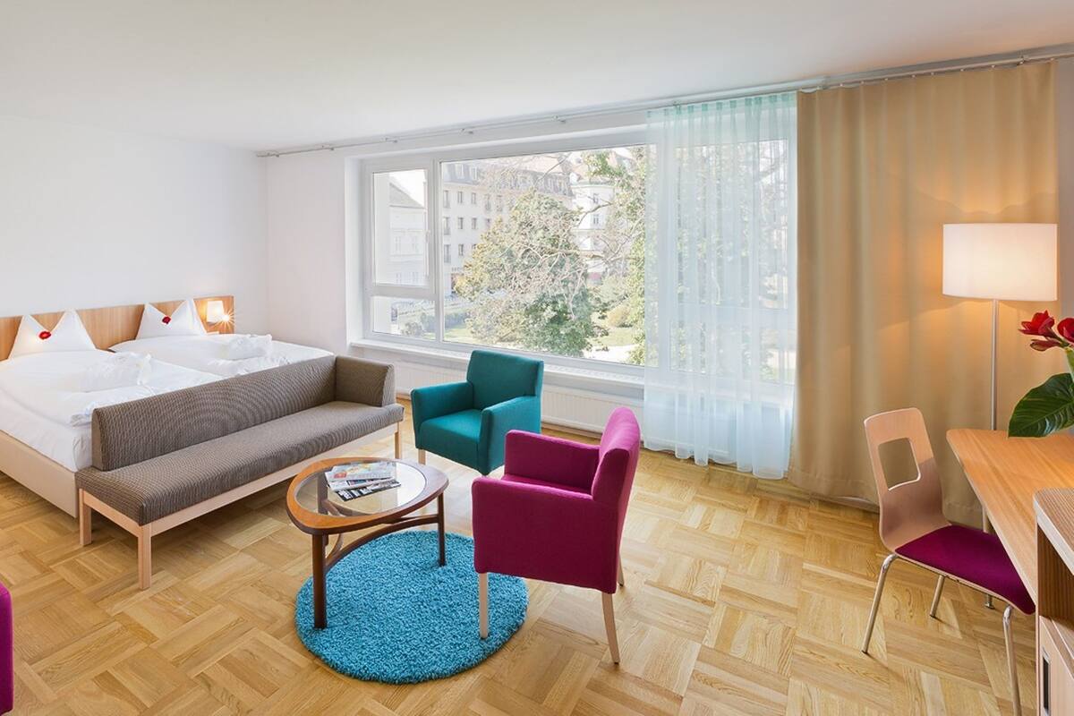 Ein Hotelzimmer mit einem Bett, einem Sofa, einem kleinen Tisch, zwei Stühlen, einer Lampe und einem Fenster mit Blick auf Gebäude und Bäume.