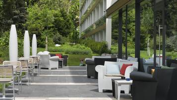 An outdoor seating area with modern furniture, surrounded by greenery and a building with large windows.
