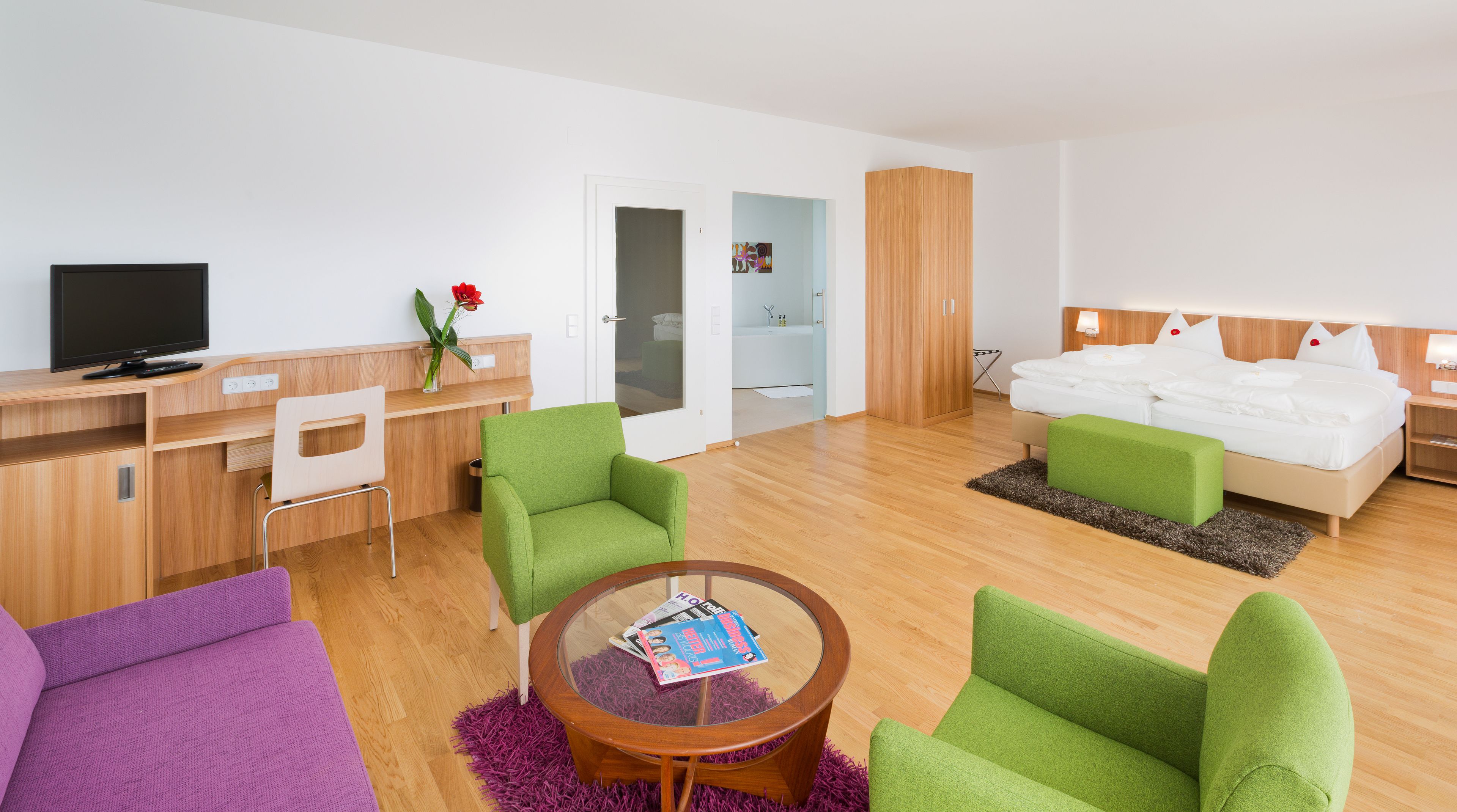 A modern hotel room with a wooden desk, green armchairs, a purple sofa, a round coffee table, and a bed with white bedding.