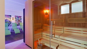 A sauna with wooden benches and a glass partition, a wooden wall with a light, and a waiting area with green chairs and artwork. - At the Park Hotel (Baden)