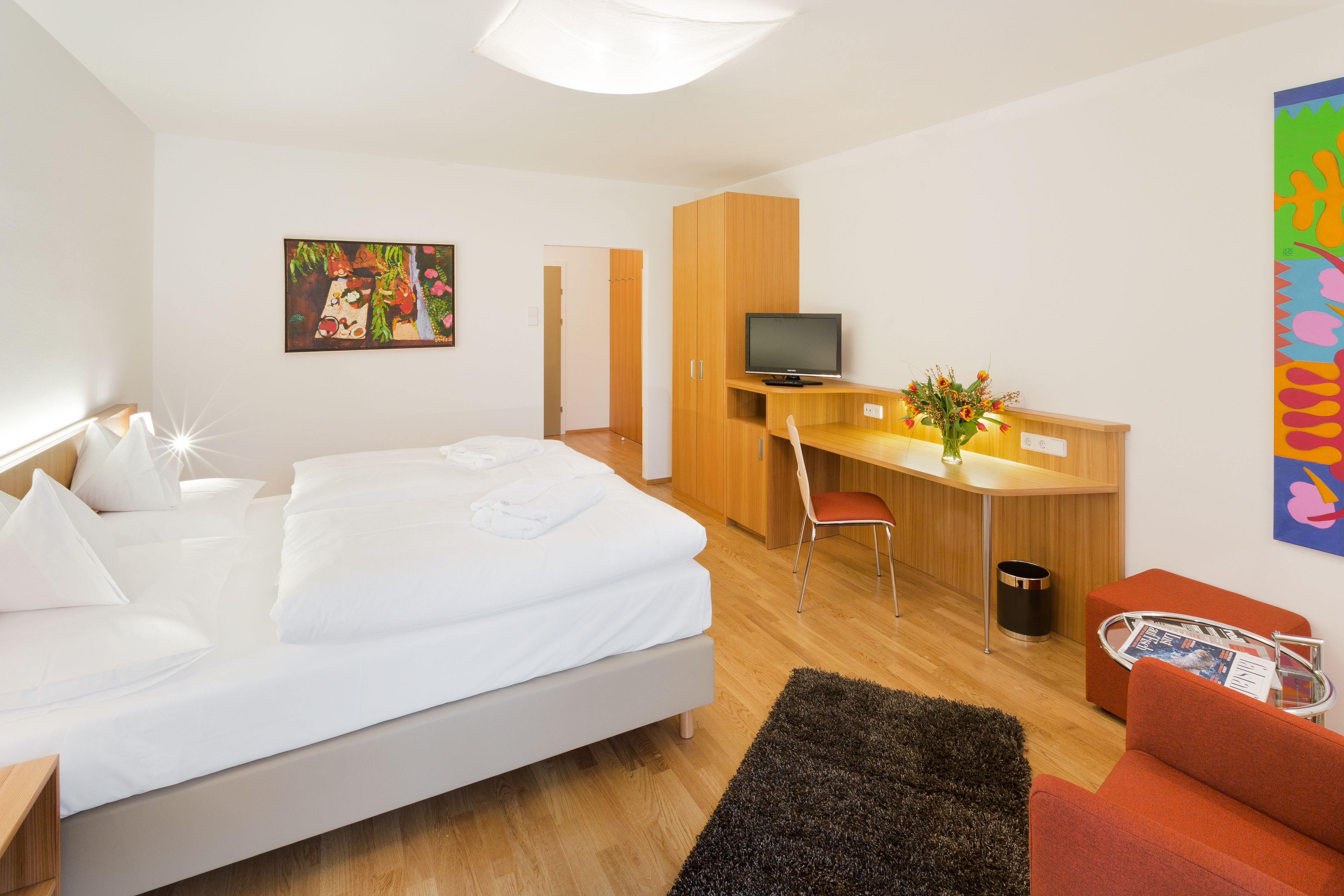 Habitación de hotel con cama, escritorio con computadora, televisor, silla, pequeña mesa con un jarrón de flores y una obra de arte de pared colorida.