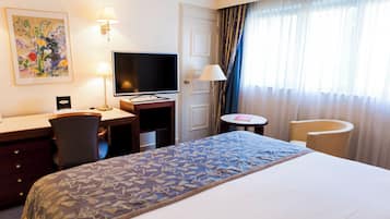 Habitación de hotel con cama, escritorio, silla, televisor, lámpara y cortinas.