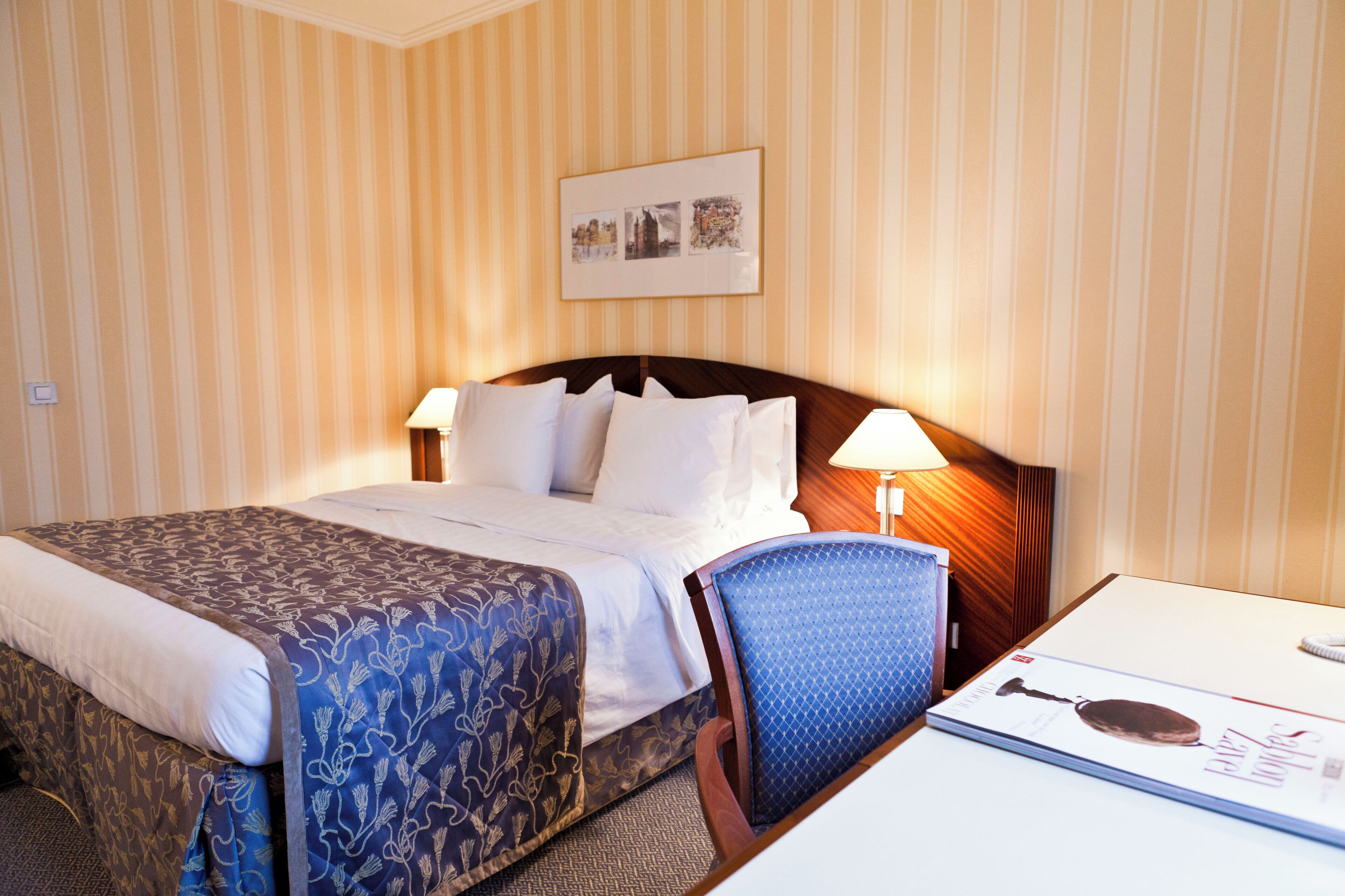 A hotel room with a bed, two bedside lamps, a chair, a desk, and a framed picture on the wall.