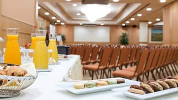 A breakfast buffet with pastries, macarons, and orange juice in a conference room with rows of chairs and a projector screen.